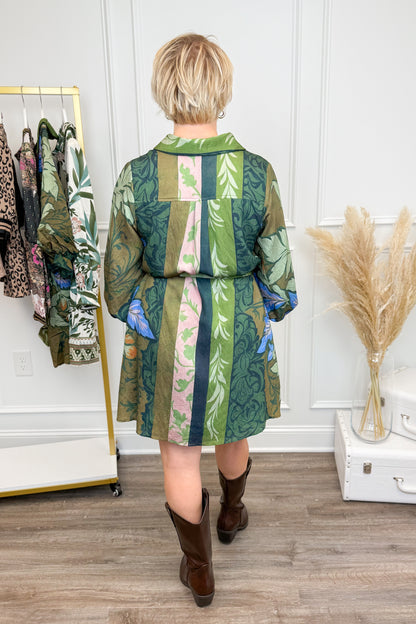 Person wearing a green and brown patterned dress, standing in a room with a rack of clothes and decorative items.