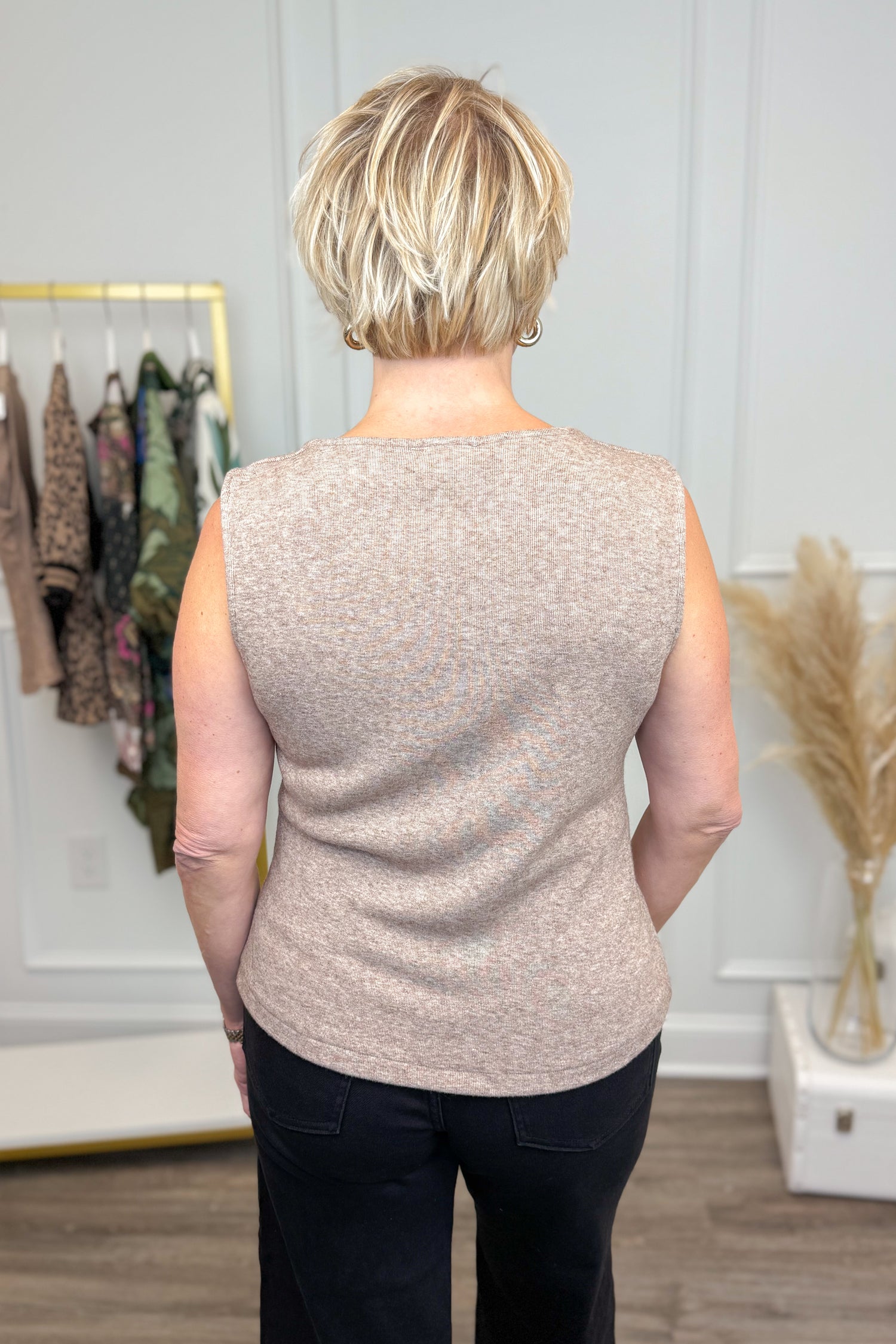 Person wearing a beige sleeveless top in a room with clothing and decor.