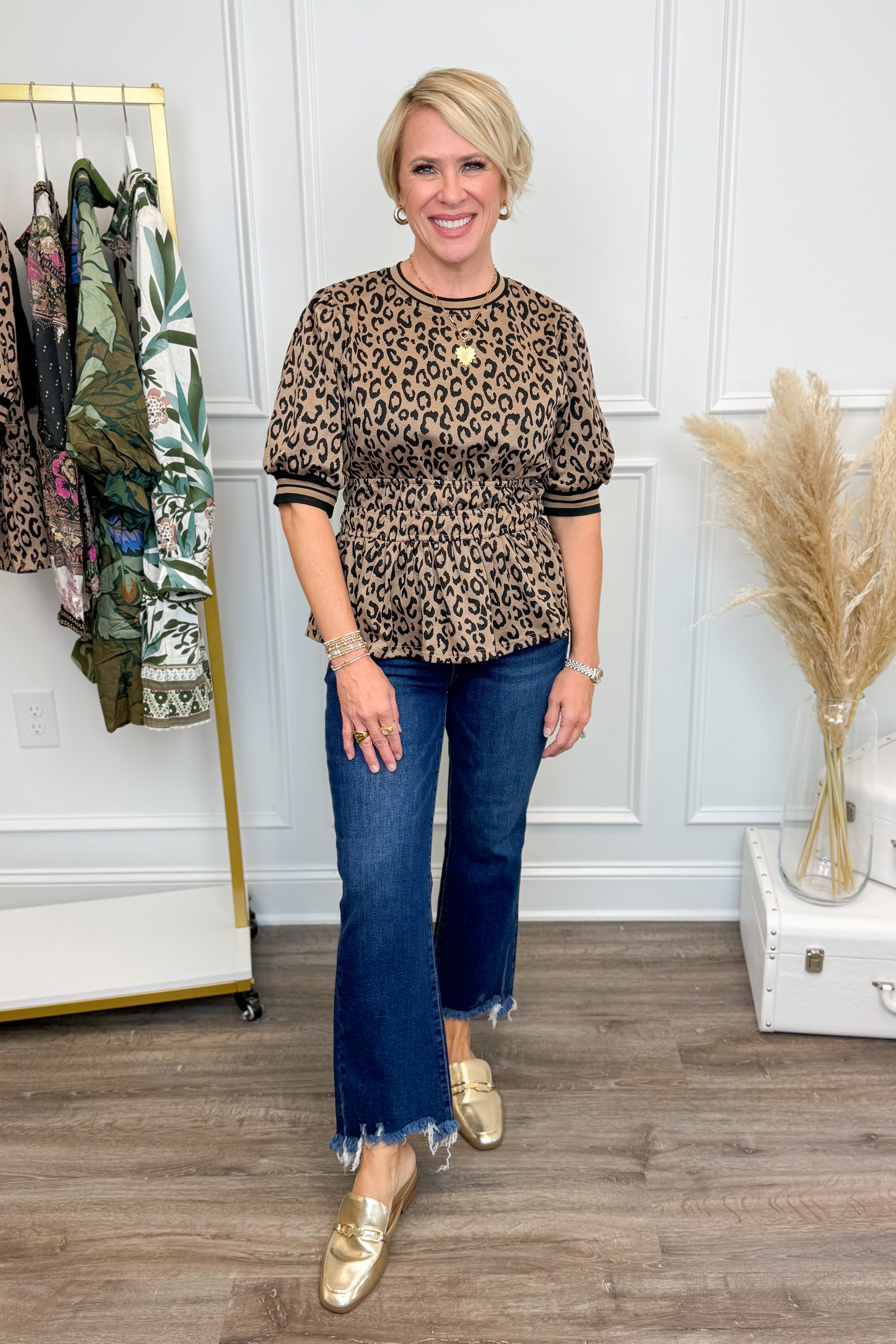 Woman wearing a leopard print top and blue jeans in a room with a clothing rack and decorative items.