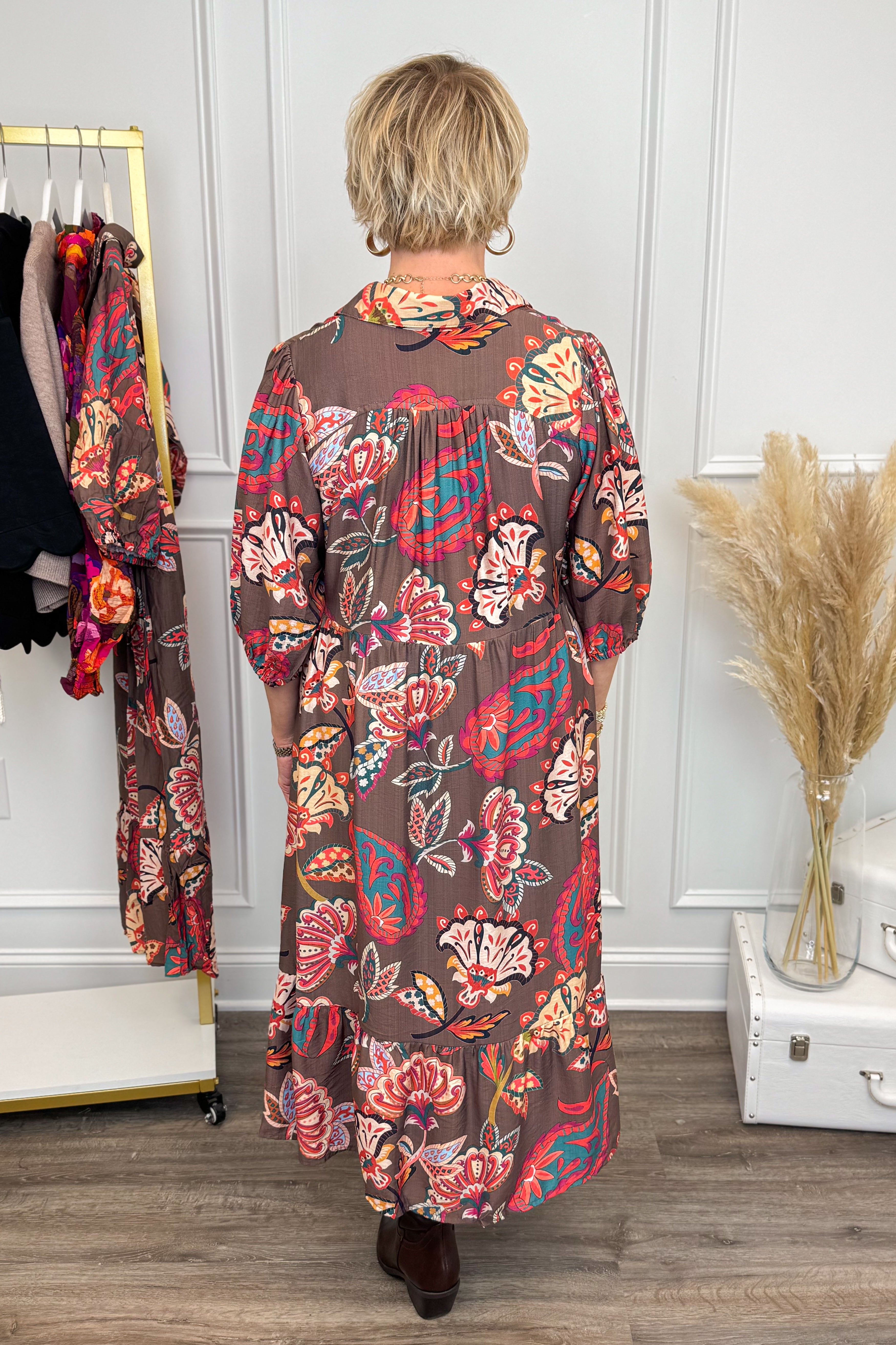 Woman wearing a long, patterned dress in a room with a white wall and wooden floor.