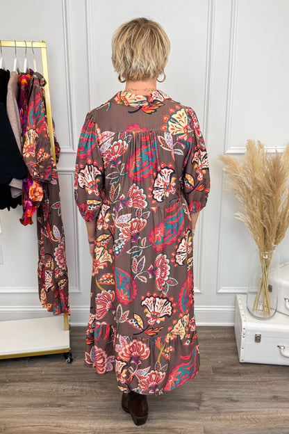 Woman wearing a long, patterned dress in a room with a white wall and wooden floor.