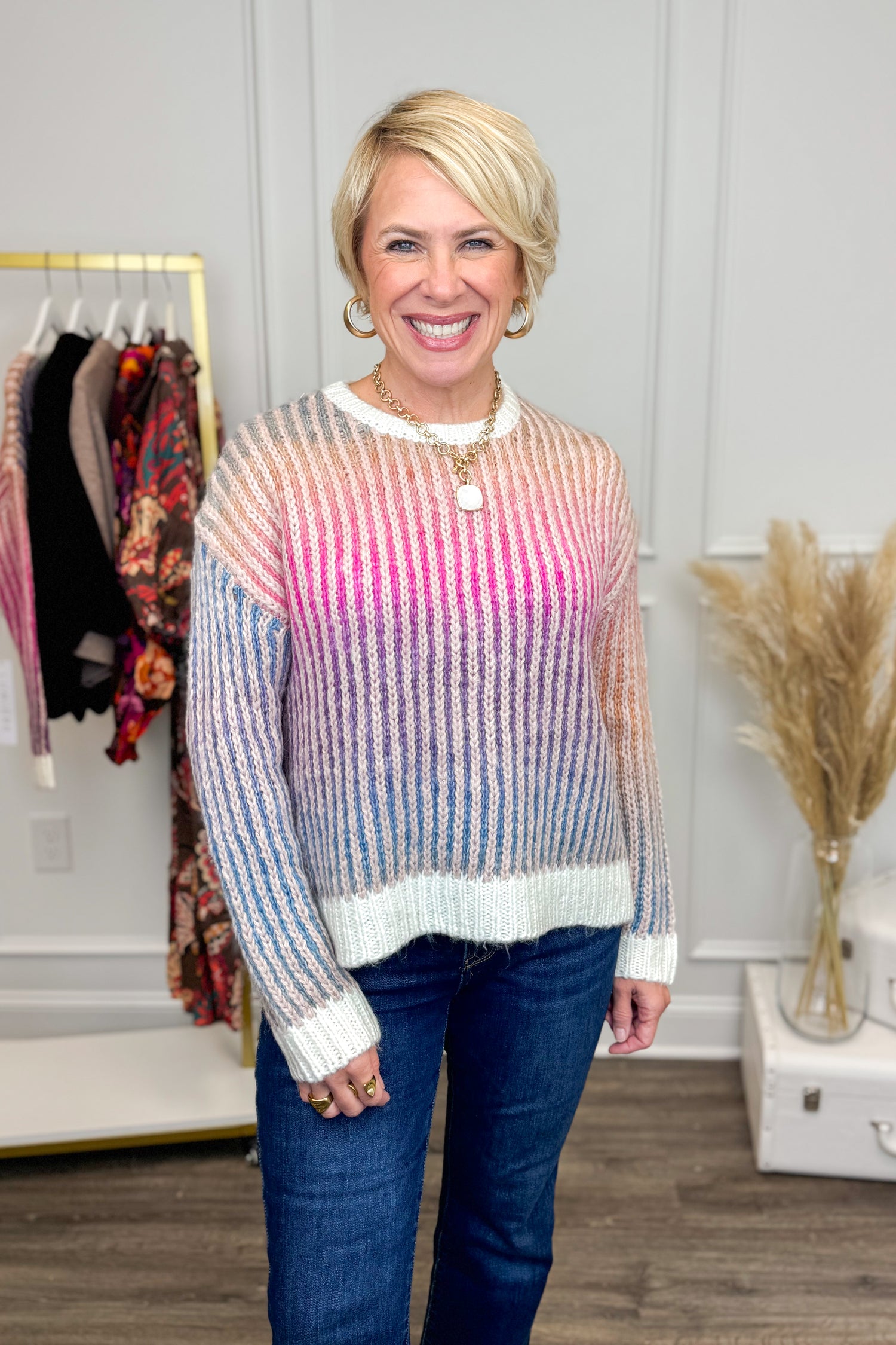 Woman wearing a colorful sweater and blue jeans in a room with clothing racks and decor.