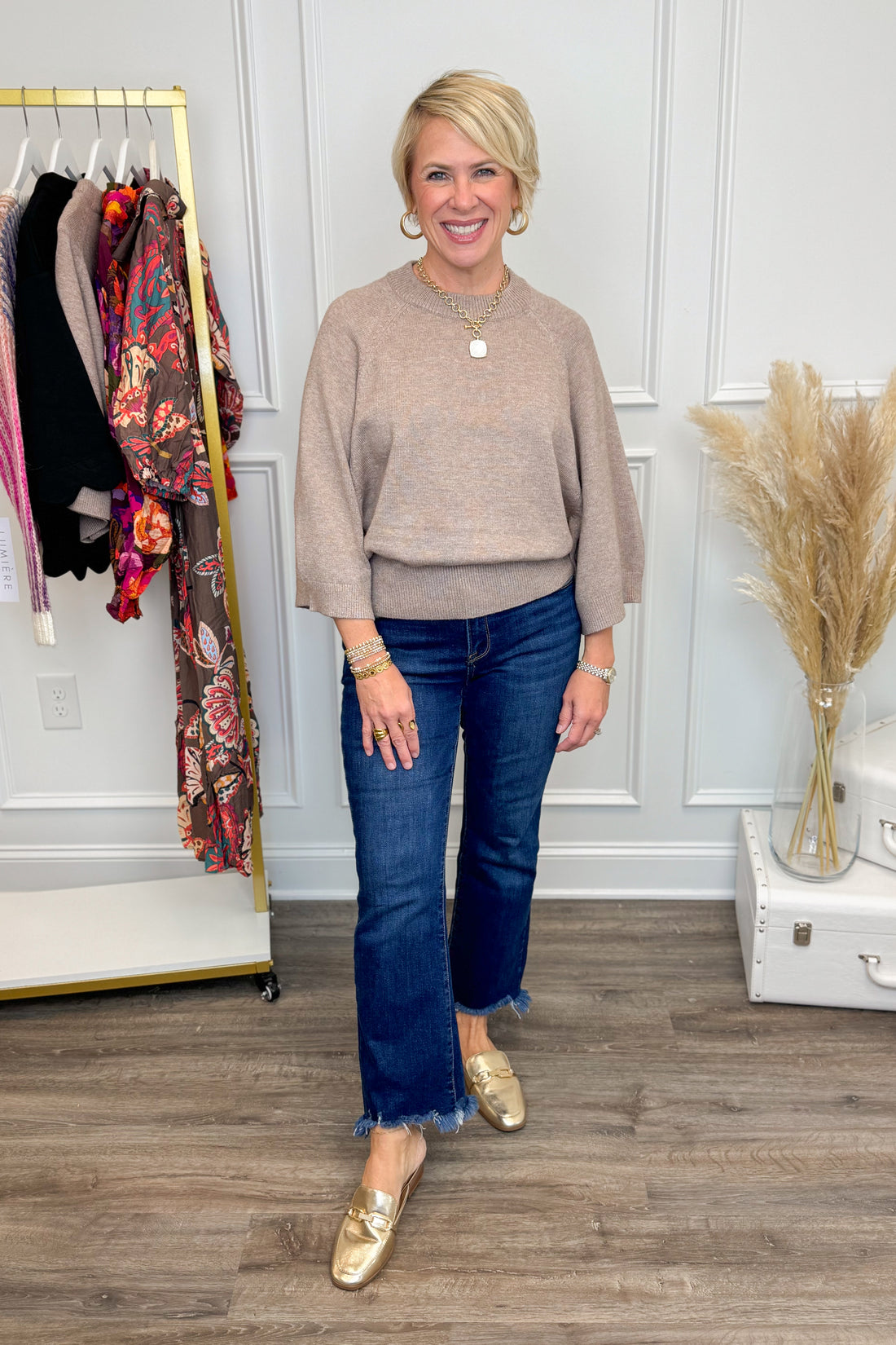 Woman wearing a beige sweater and blue jeans standing in a room with a rack of clothes and decorative elements.