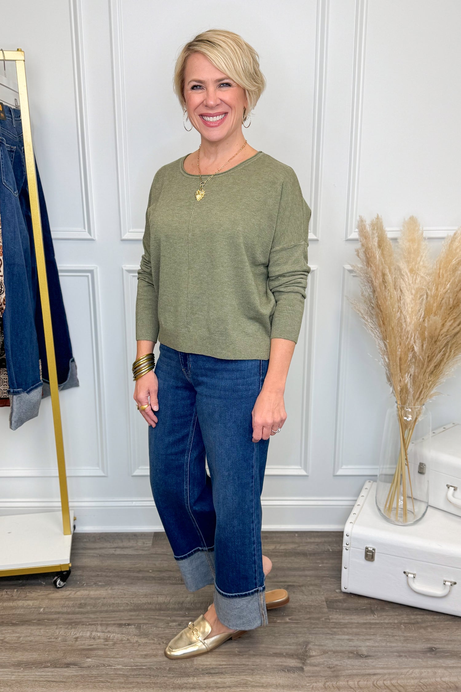 Woman wearing a green sweater and blue jeans standing in a room with white paneled walls and decorative elements.