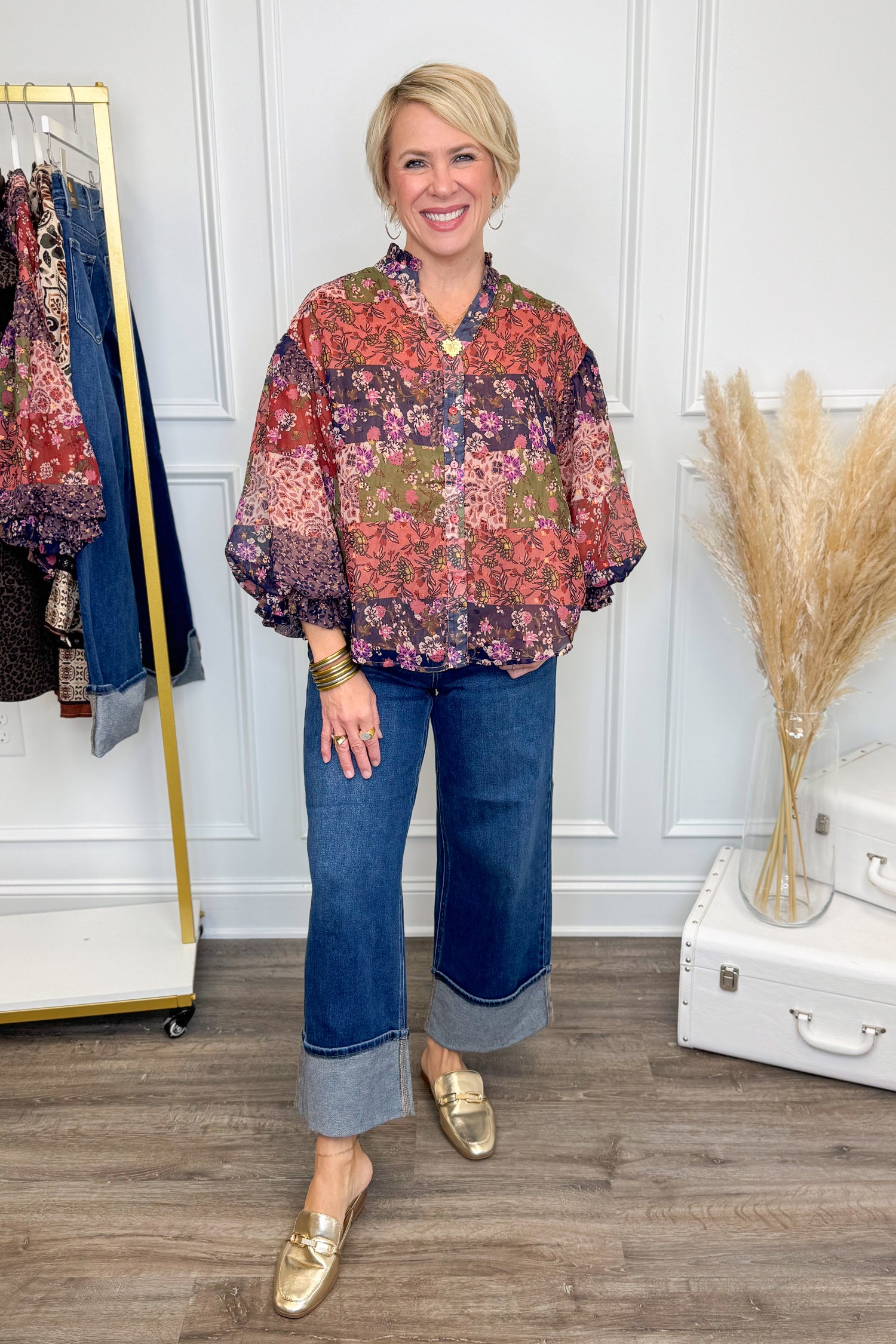 Woman wearing a patterned blouse and blue jeans standing in a room with a white wall and wooden floor.