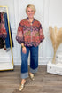 Woman wearing a patterned blouse and blue jeans standing in a room with a white wall and wooden floor.