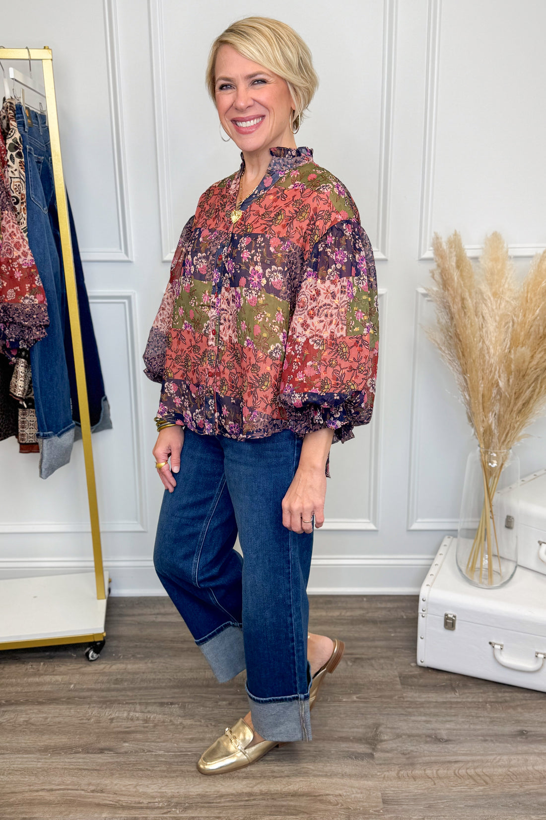Woman wearing a patterned blouse and jeans standing in a room with a wardrobe and decorative elements.