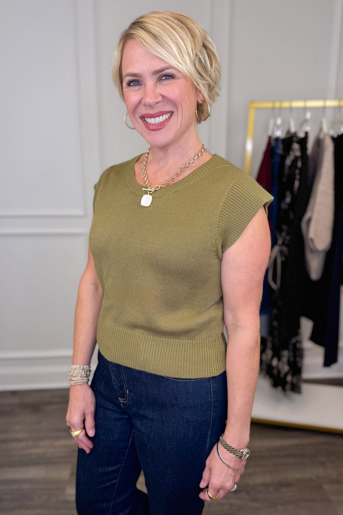 Woman wearing a green sleeveless sweater and dark pants in a clothing store.