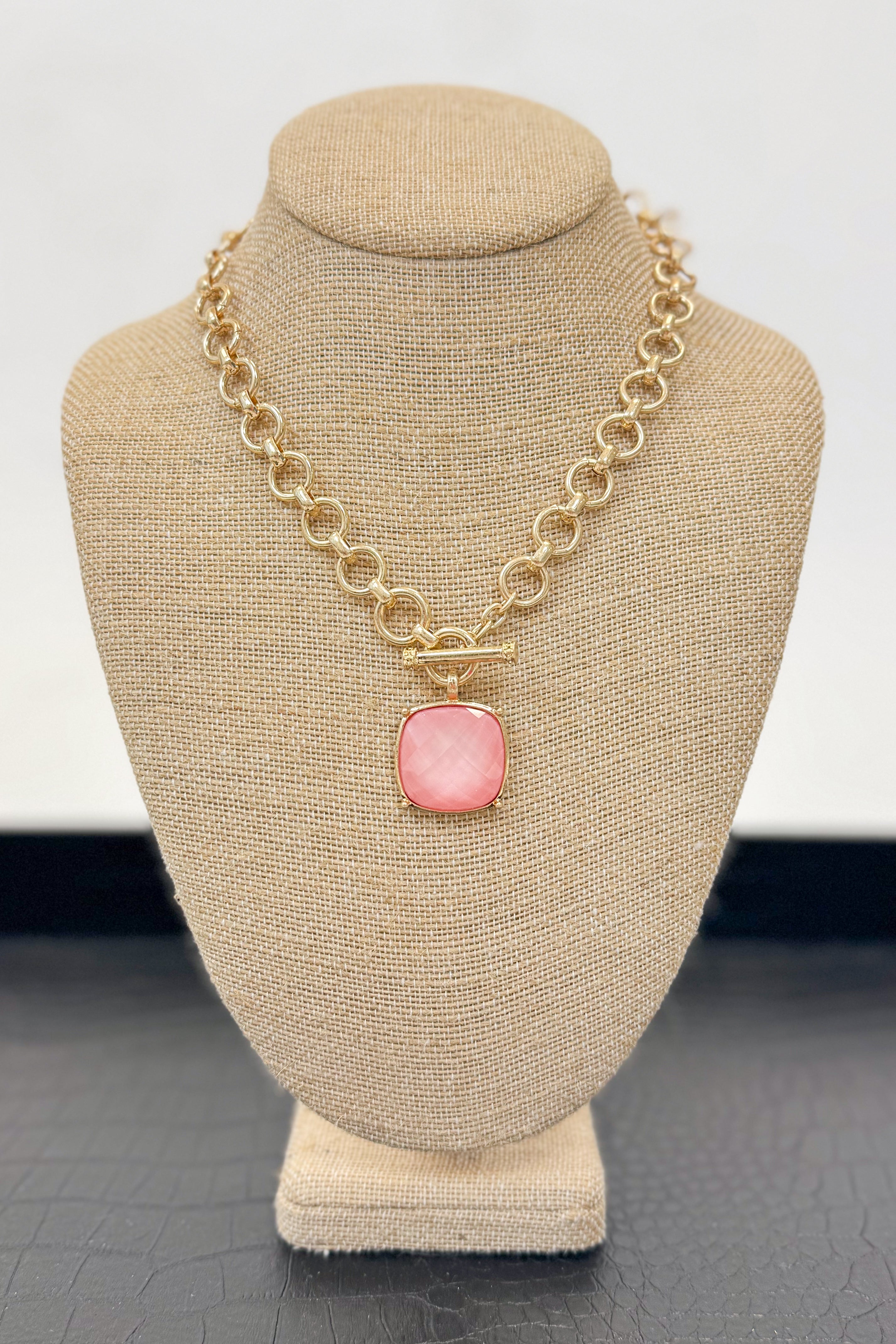 Gold necklace with a light pink pendant on a beige bust against a white background