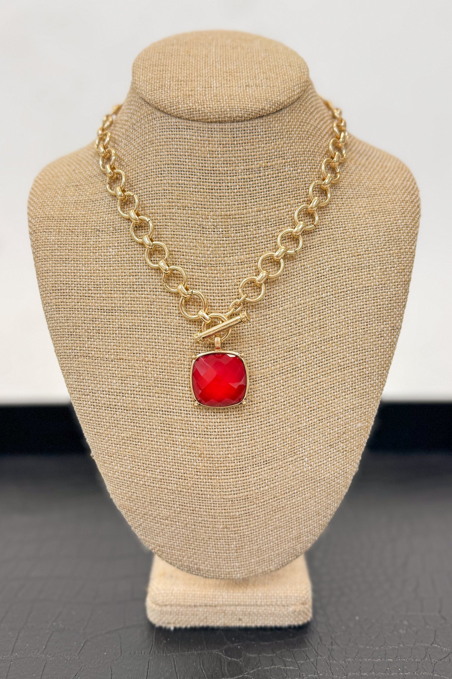 Gold necklace with a red pendant on a beige bust against a white background