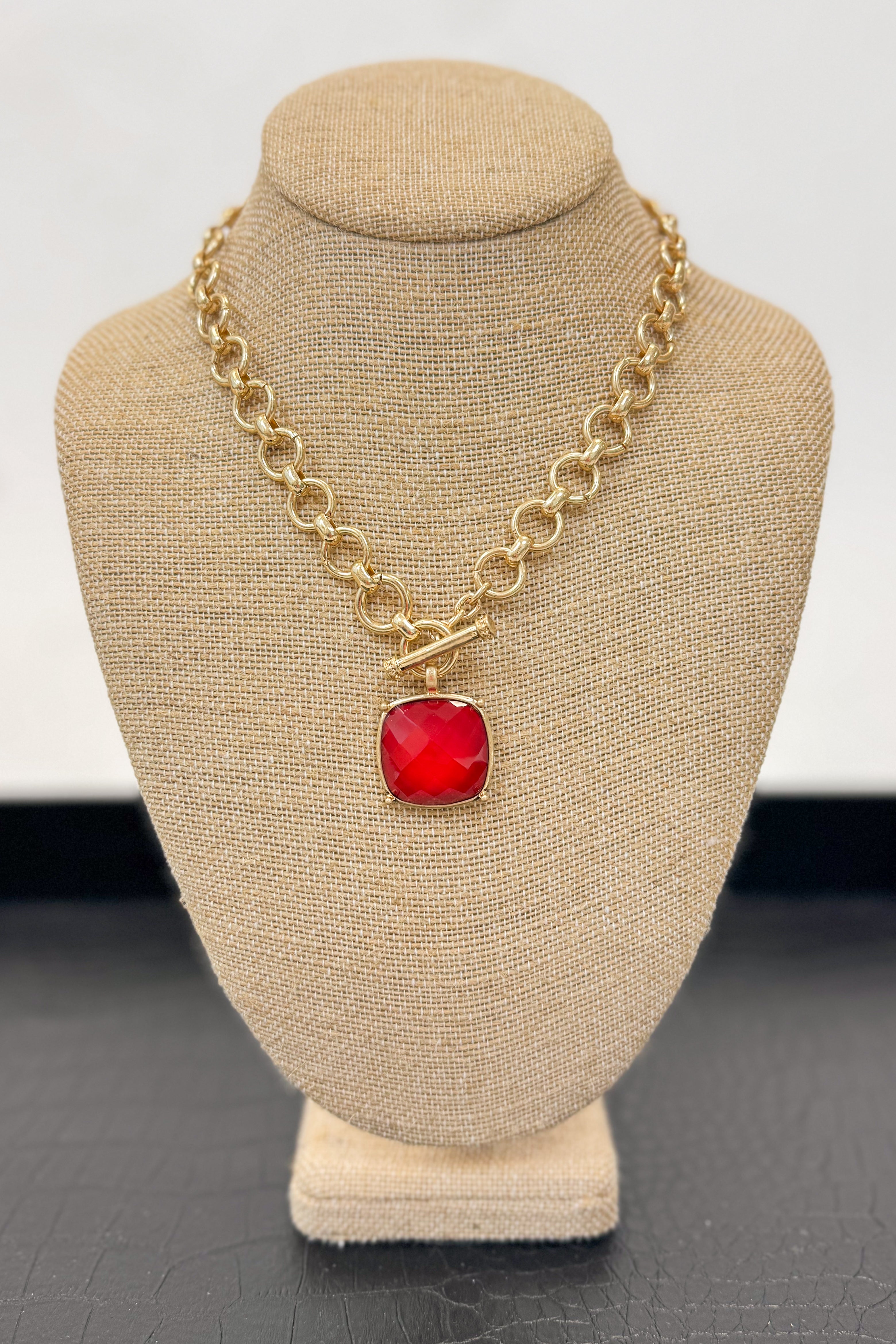 Gold necklace with a red pendant on a beige bust against a white background