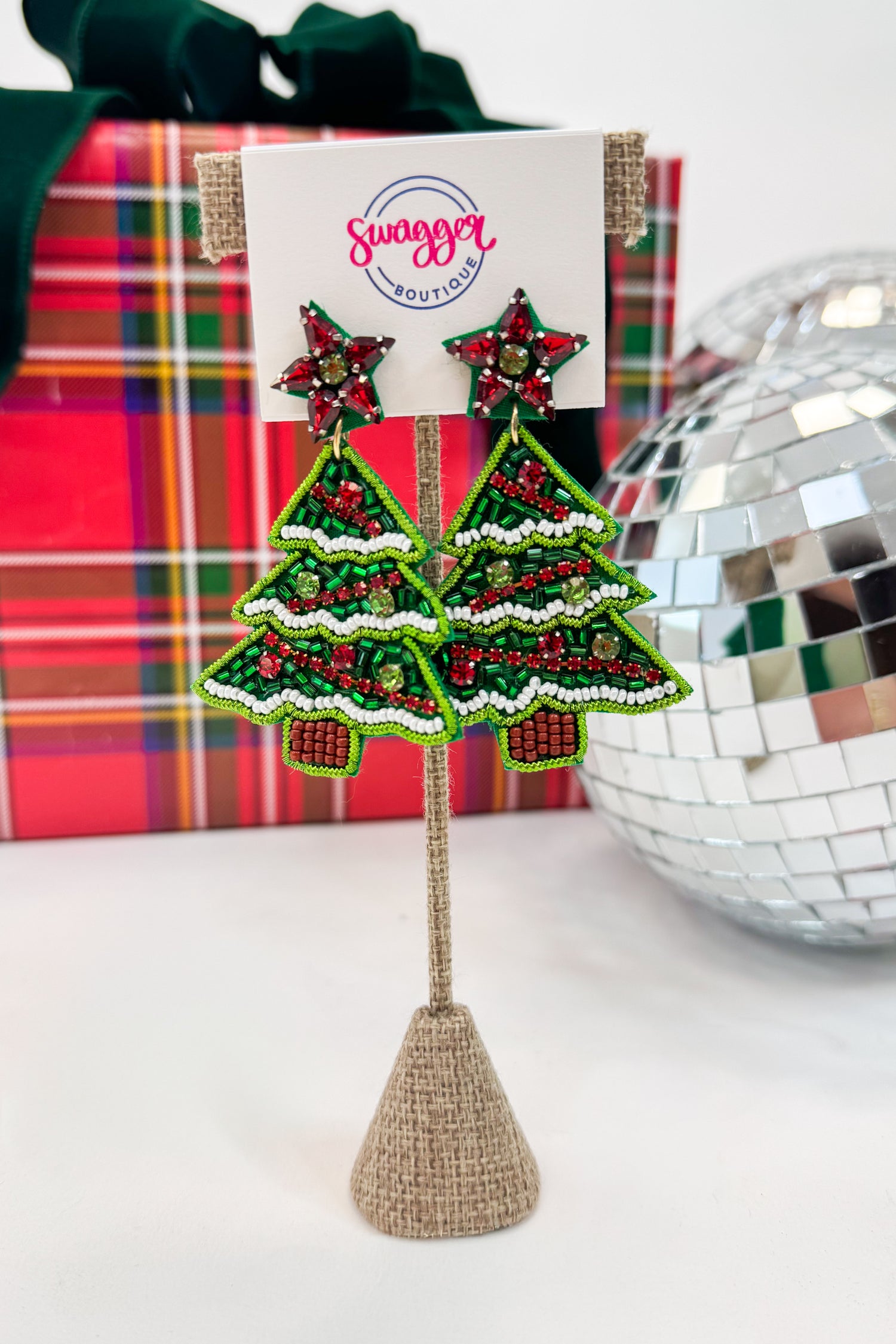 Decorative Christmas tree earrings on a stand with a gift box and disco ball in the background