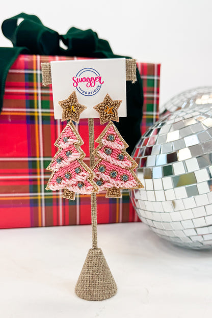 Decorative earrings shaped like Christmas trees on a stand with a gift box and disco ball in the background.