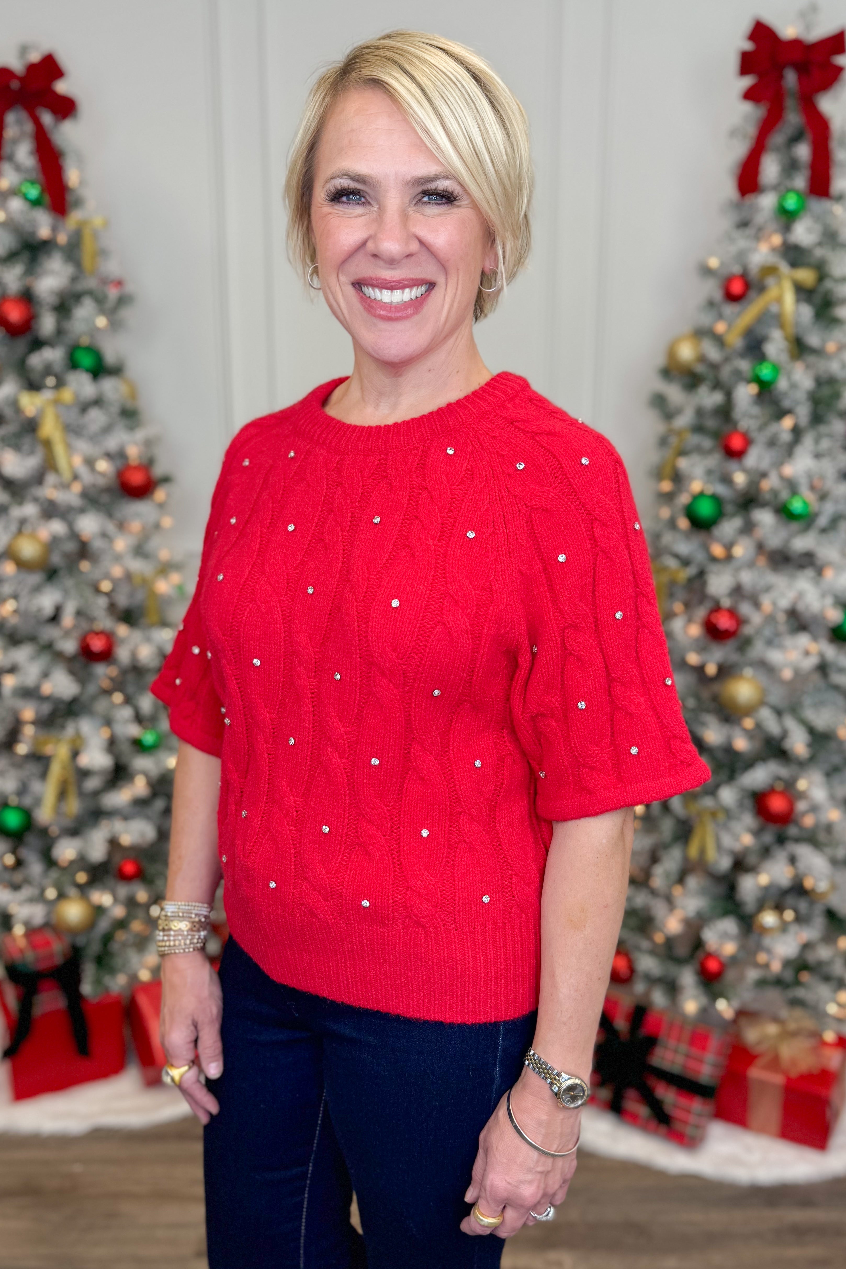Woman wearing a red sweater with rhinestone in front of Christmas trees.