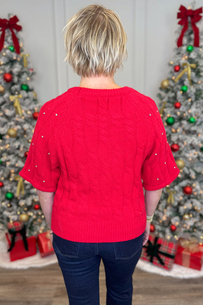 Woman wearing a red sweater with rhinestone in front of Christmas trees.