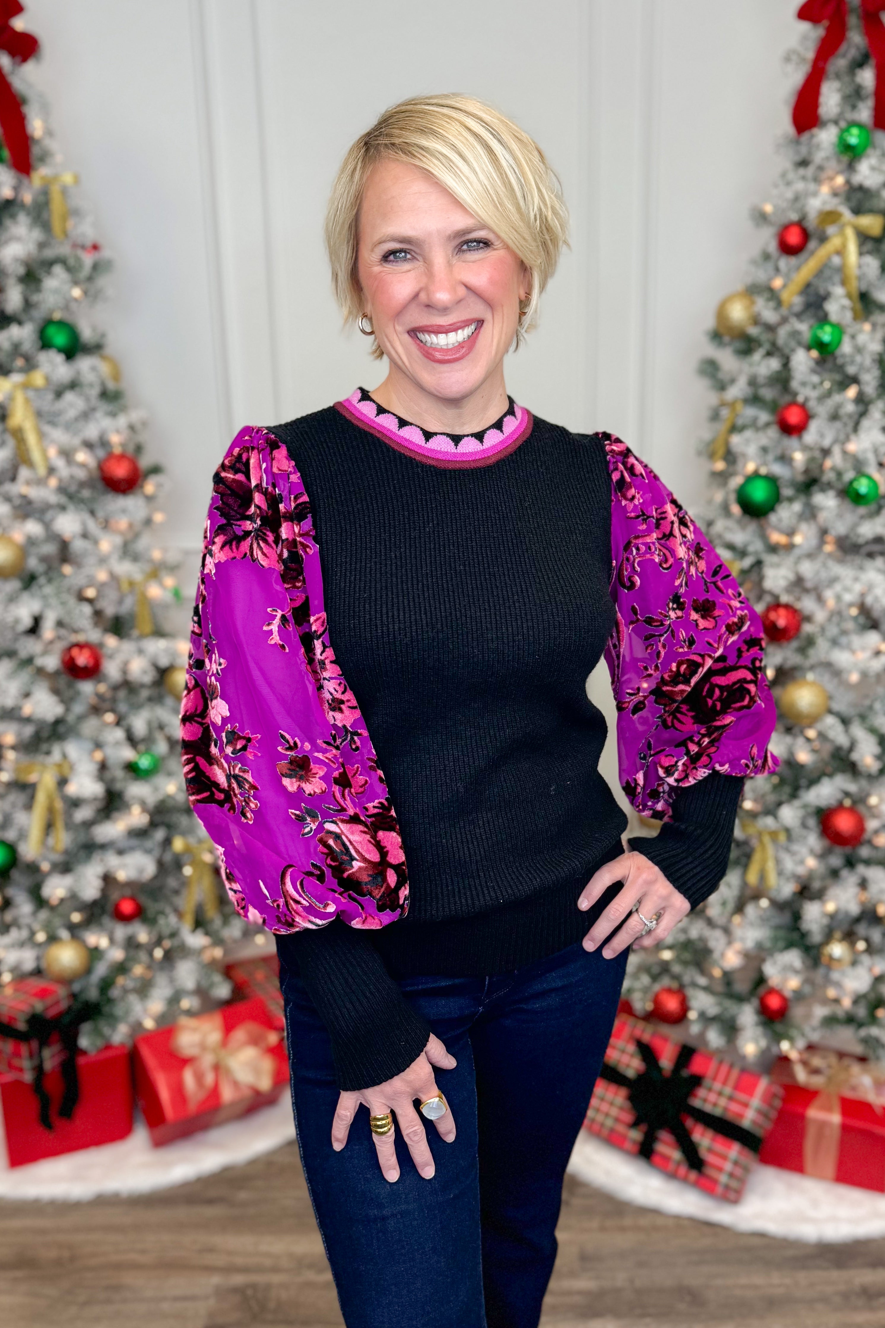 Woman wearing a black sweater with a colorful floral jacket in front of Christmas trees.