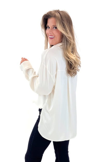 Side view of woman wearing Cream Oversized Satin Shirt with jeans against tan studio background.
