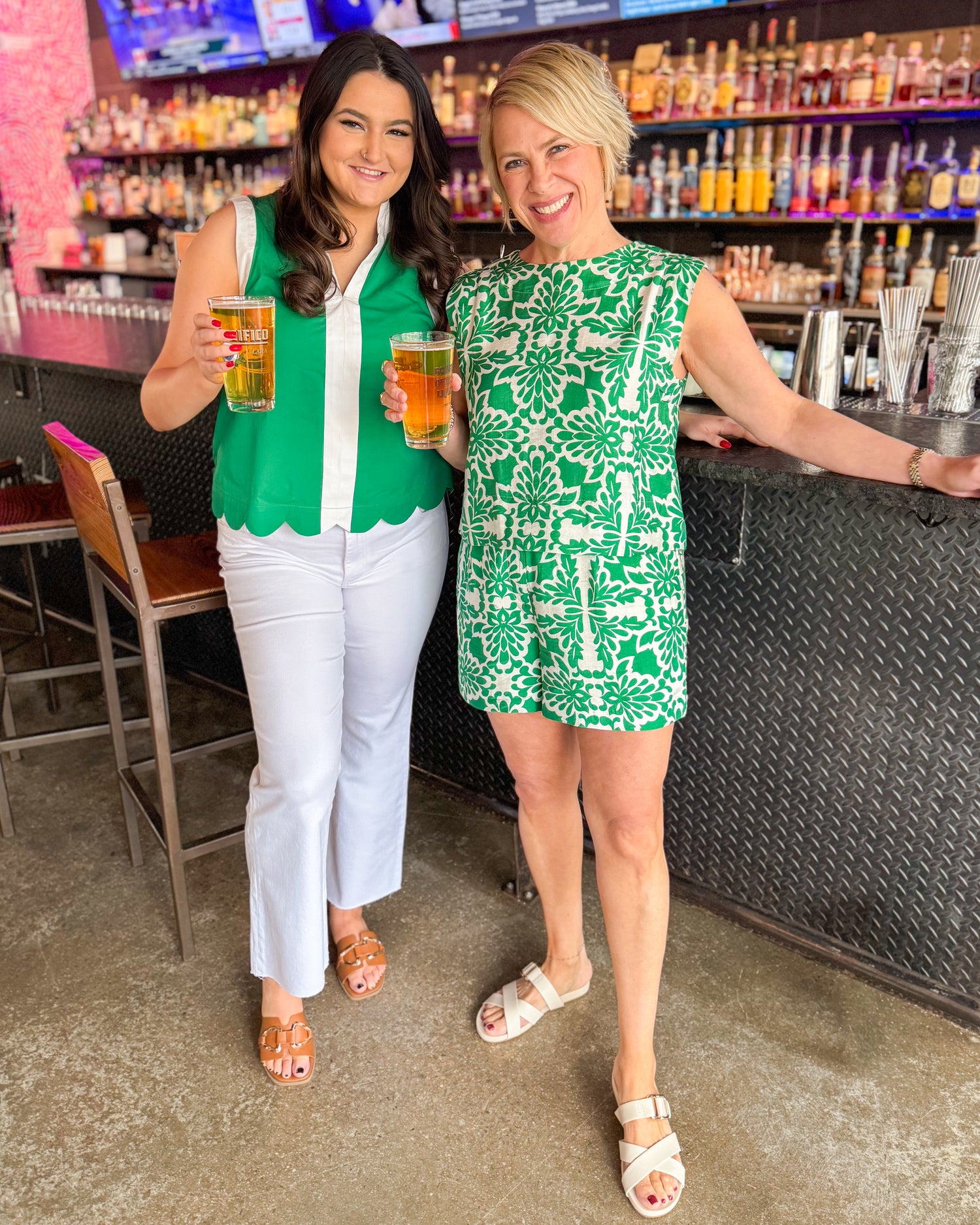 Two women in green and white outfits holding drinks in a bar setting.