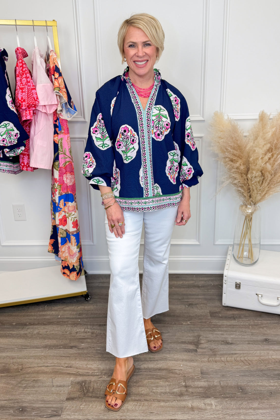 Woman wearing a colorful kimono-style top and white pants in a room with floral dresses on a rack.