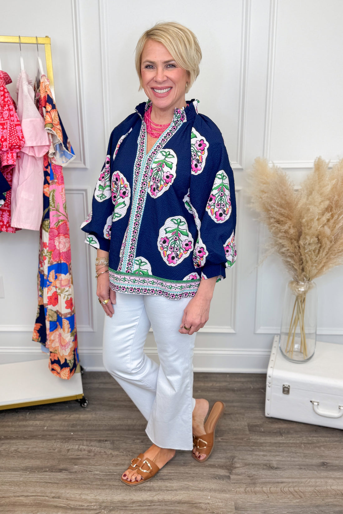 Woman wearing a colorful floral jacket in a room with a rack of clothes and decorative items.