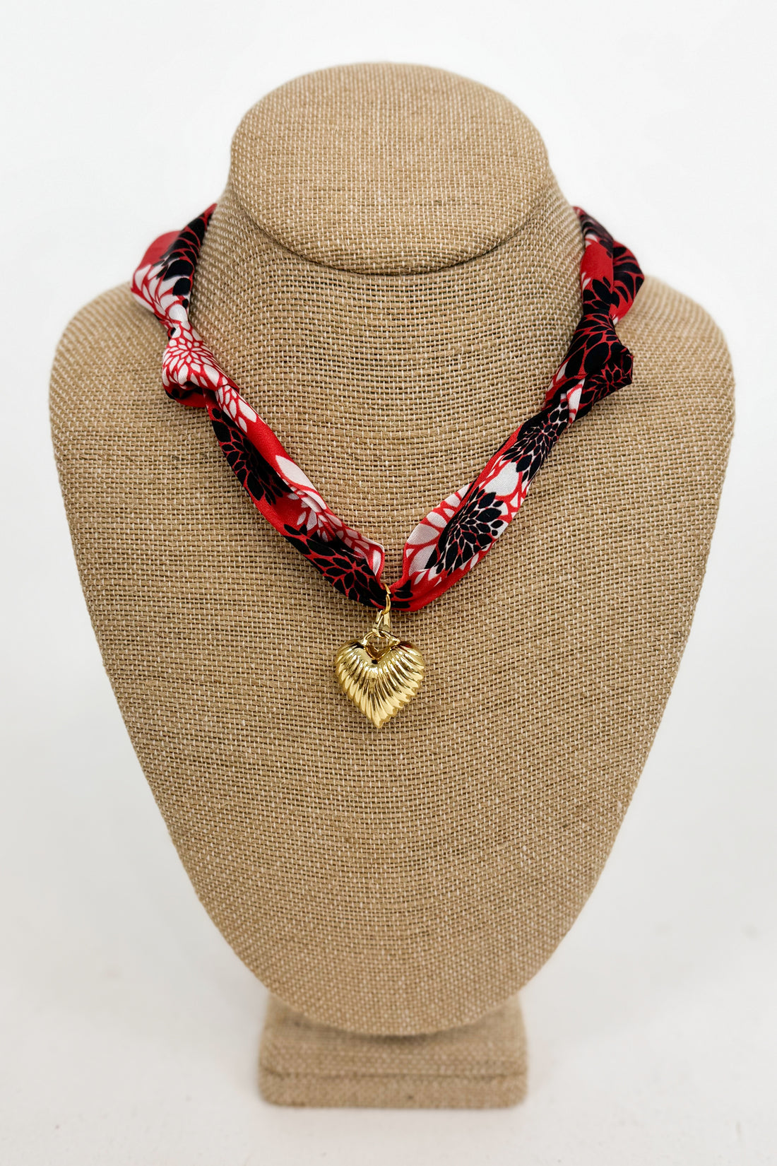 Necklace with a red and black patterned strap and gold heart pendant on a beige bust.