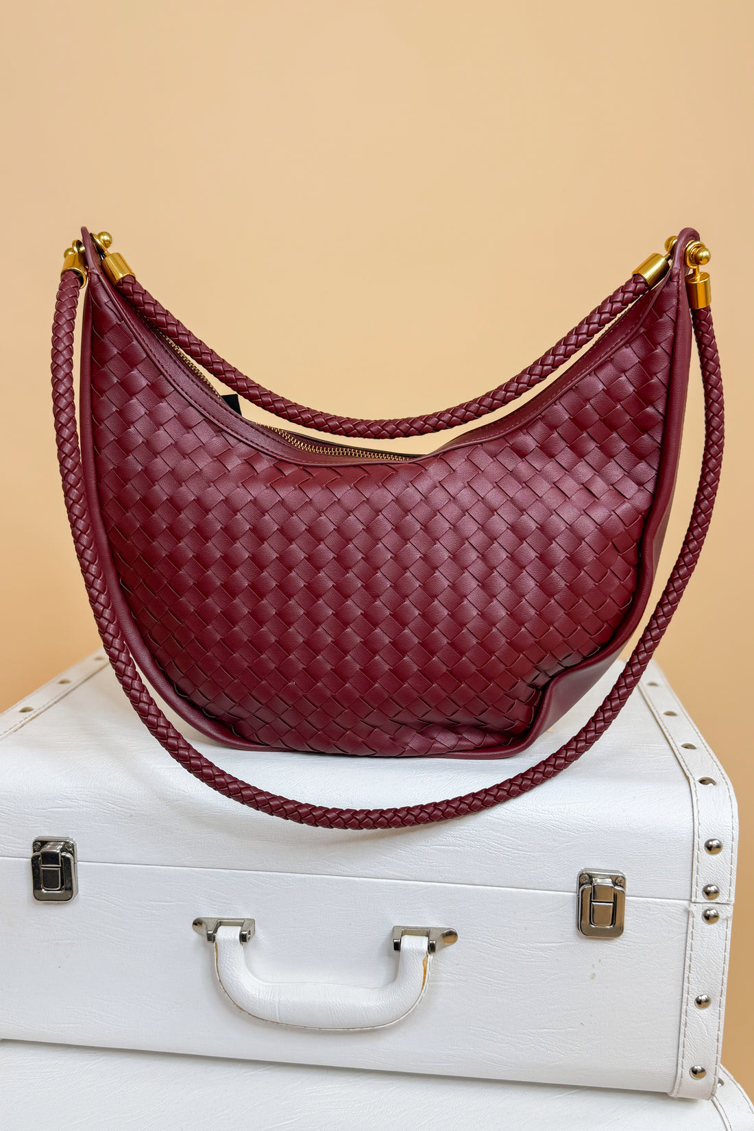 Maroon woven handbag with gold accents on a white suitcase against a beige background