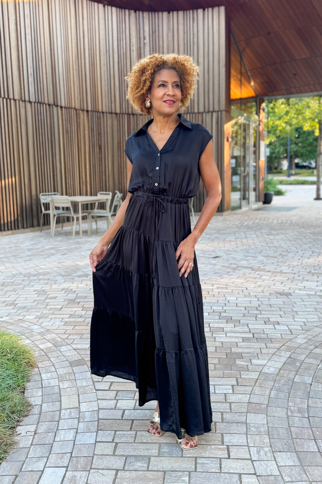 Woman in a black dress standing outdoors on a paved area with wooden walls in the background.