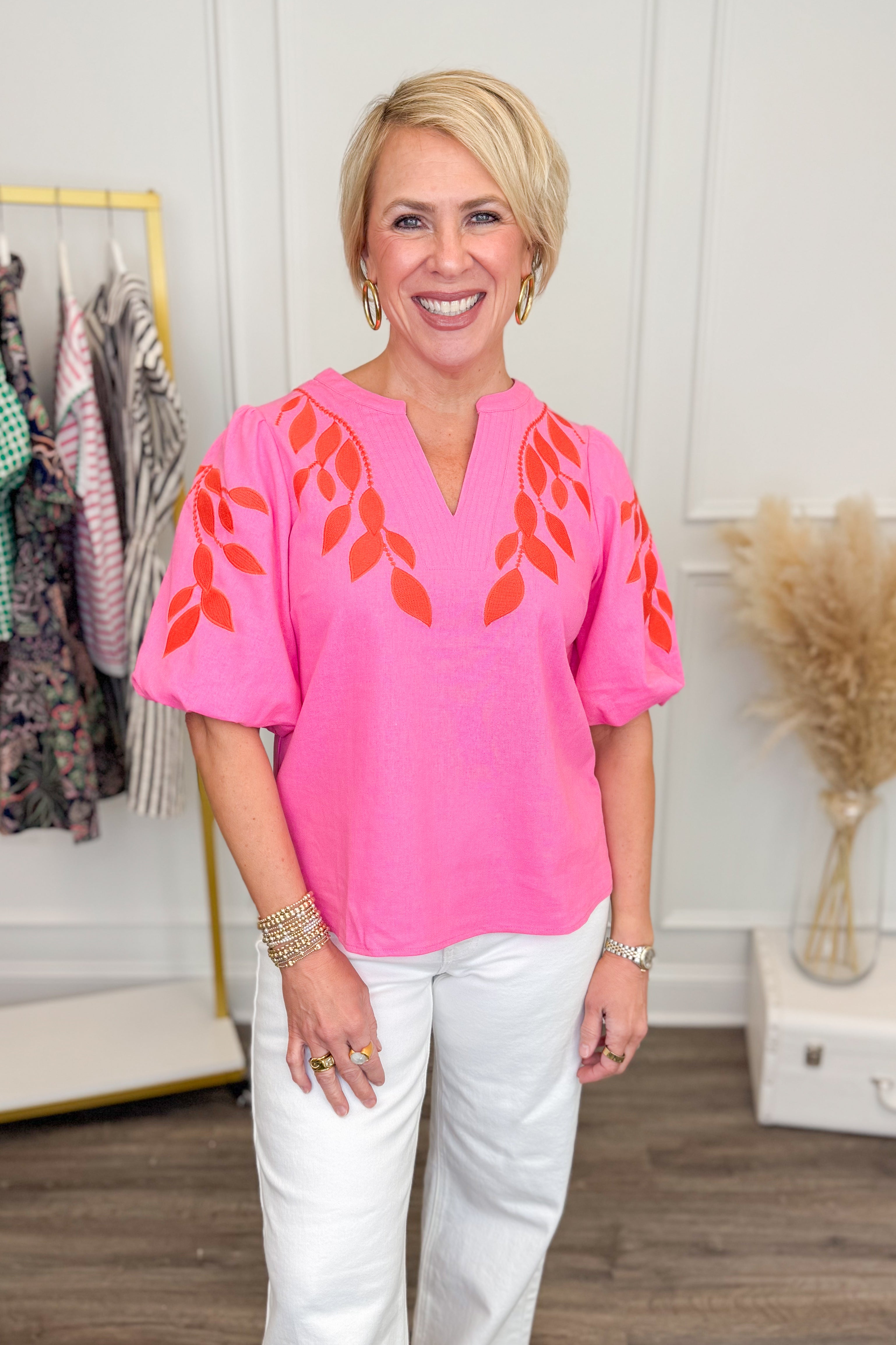 Woman wearing Pink &amp; Orange Floral Embroidered V-Neck Top with jeans with clothes &amp; decor in the background.