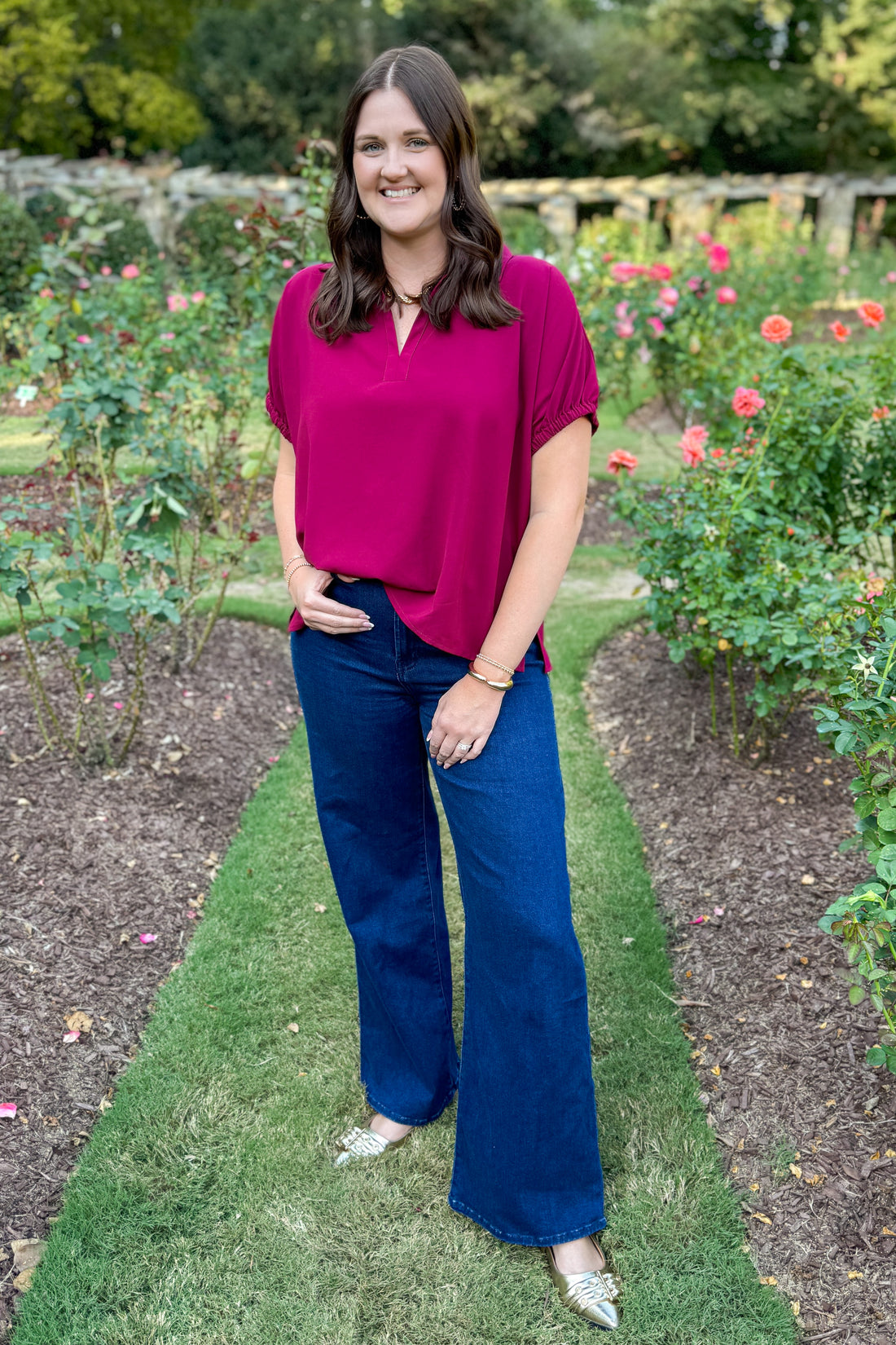 Woman in a pink top and blue pants standing in a garden with flowers and greenery.