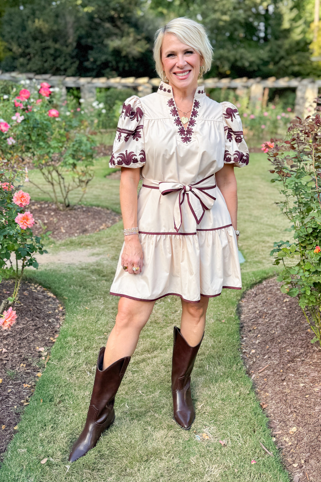 Woman in a white dress with red patterns and brown boots standing in a garden.