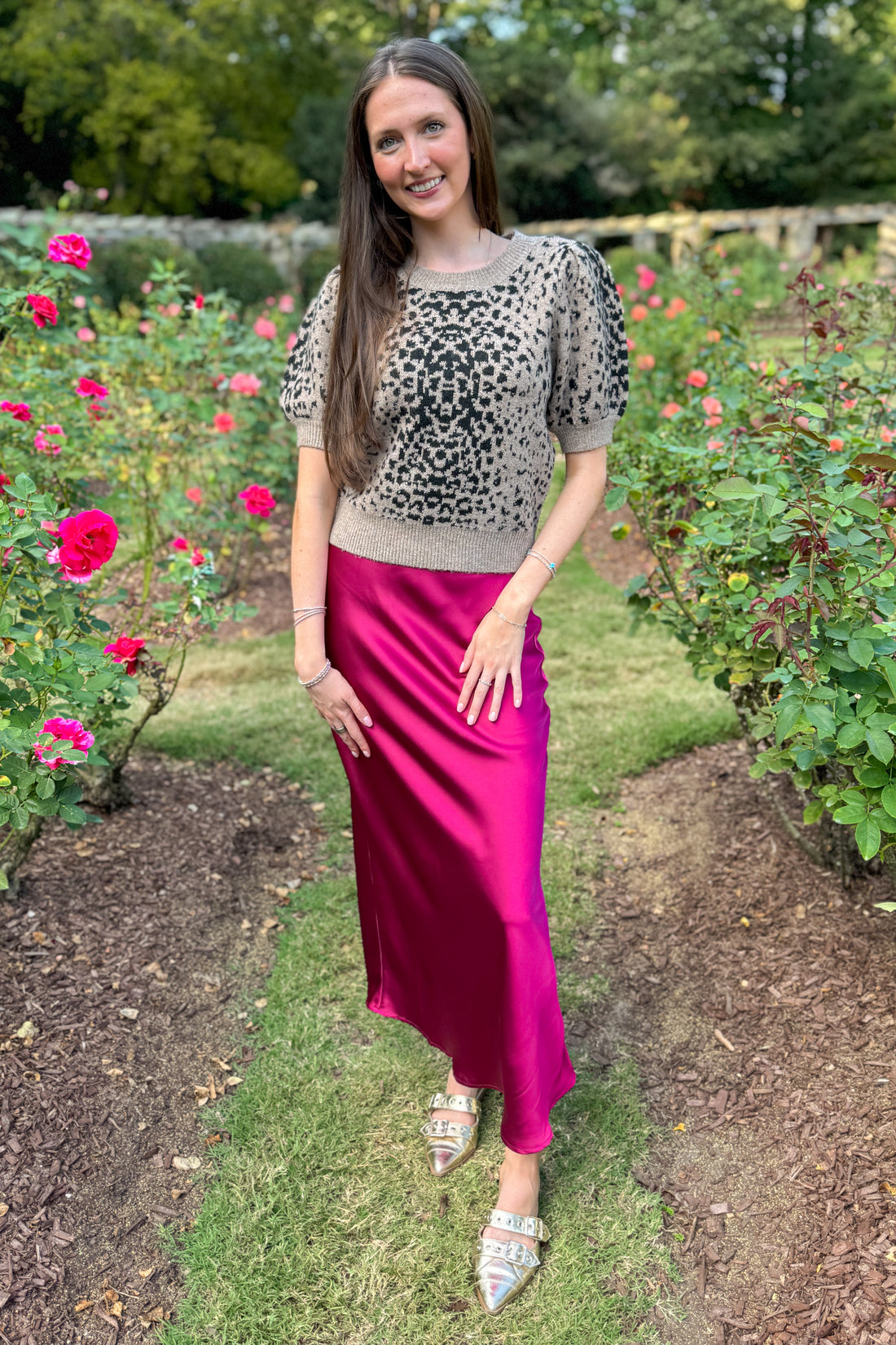Woman in a leopard print top and pink skirt standing in a garden with flowers.