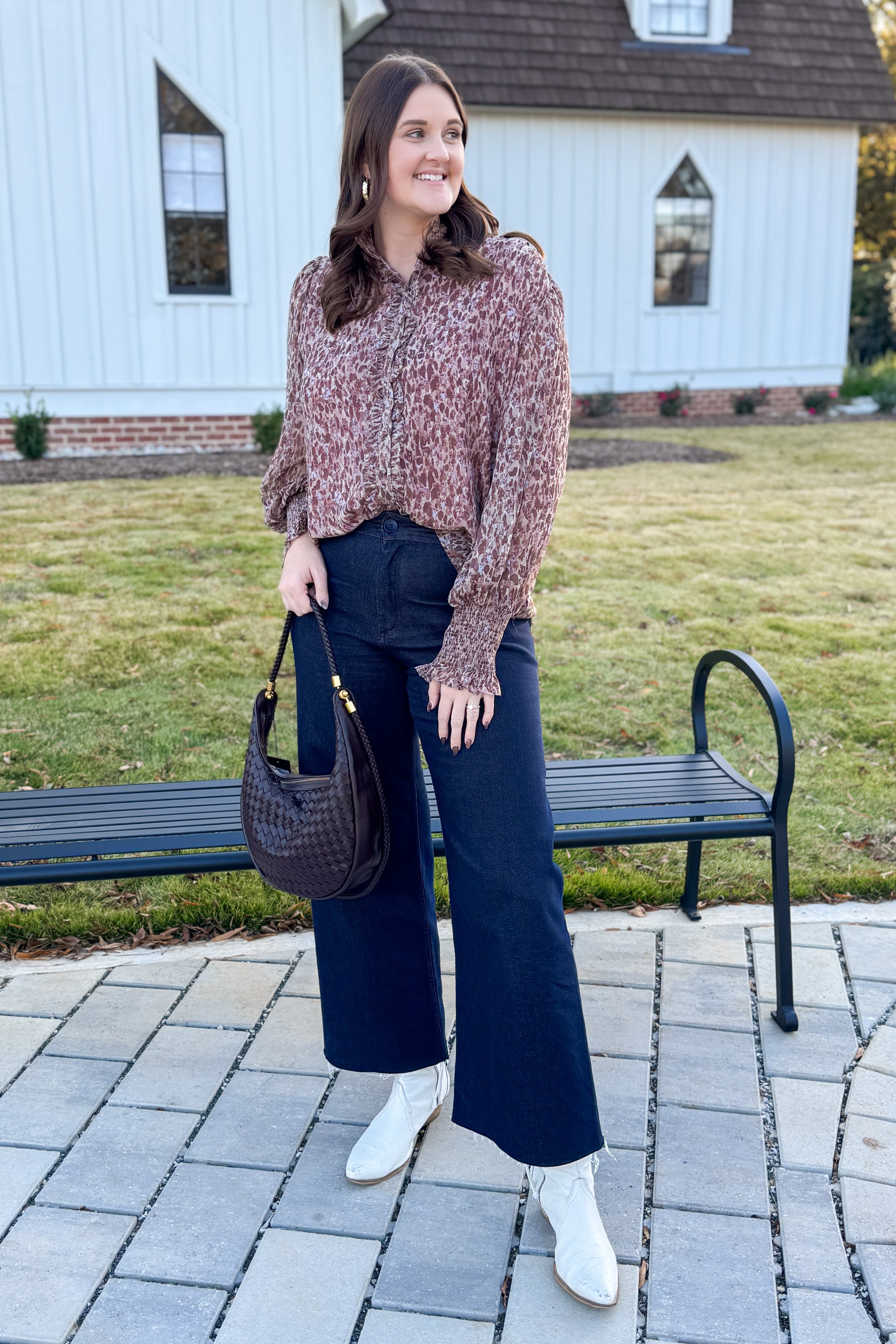 Woman standing outdoors in front of a white building, wearing a patterned sweater, dark blue jeans, and white boots.