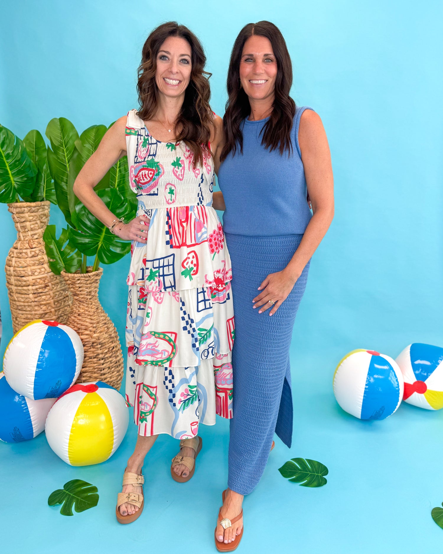 Two women standing next to colorful beach balls and a plant against a blue background