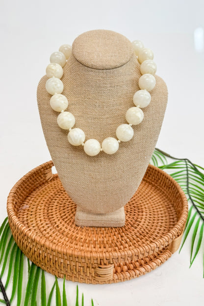Beige necklace on a mannequin bust with a woven base, surrounded by green leaves.