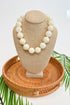 Beige necklace on a mannequin bust with a woven base, surrounded by green leaves.