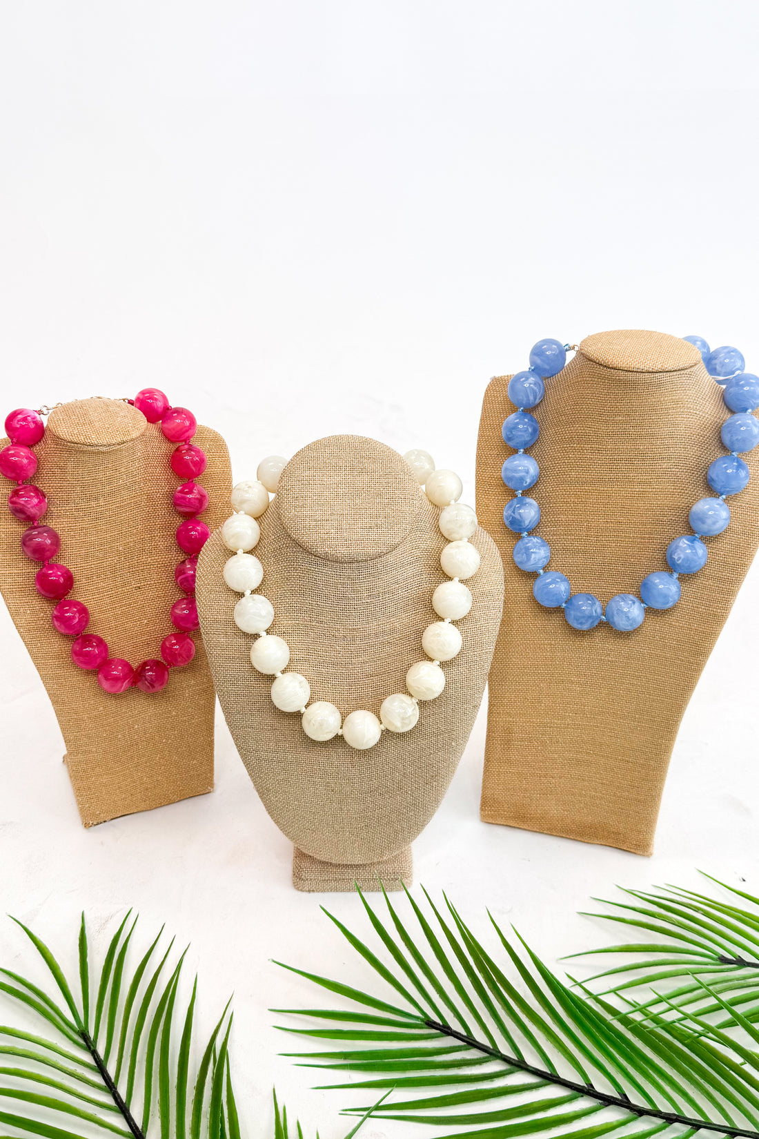 Three necklaces on mannequin heads with a white background and green leaves at the bottom.