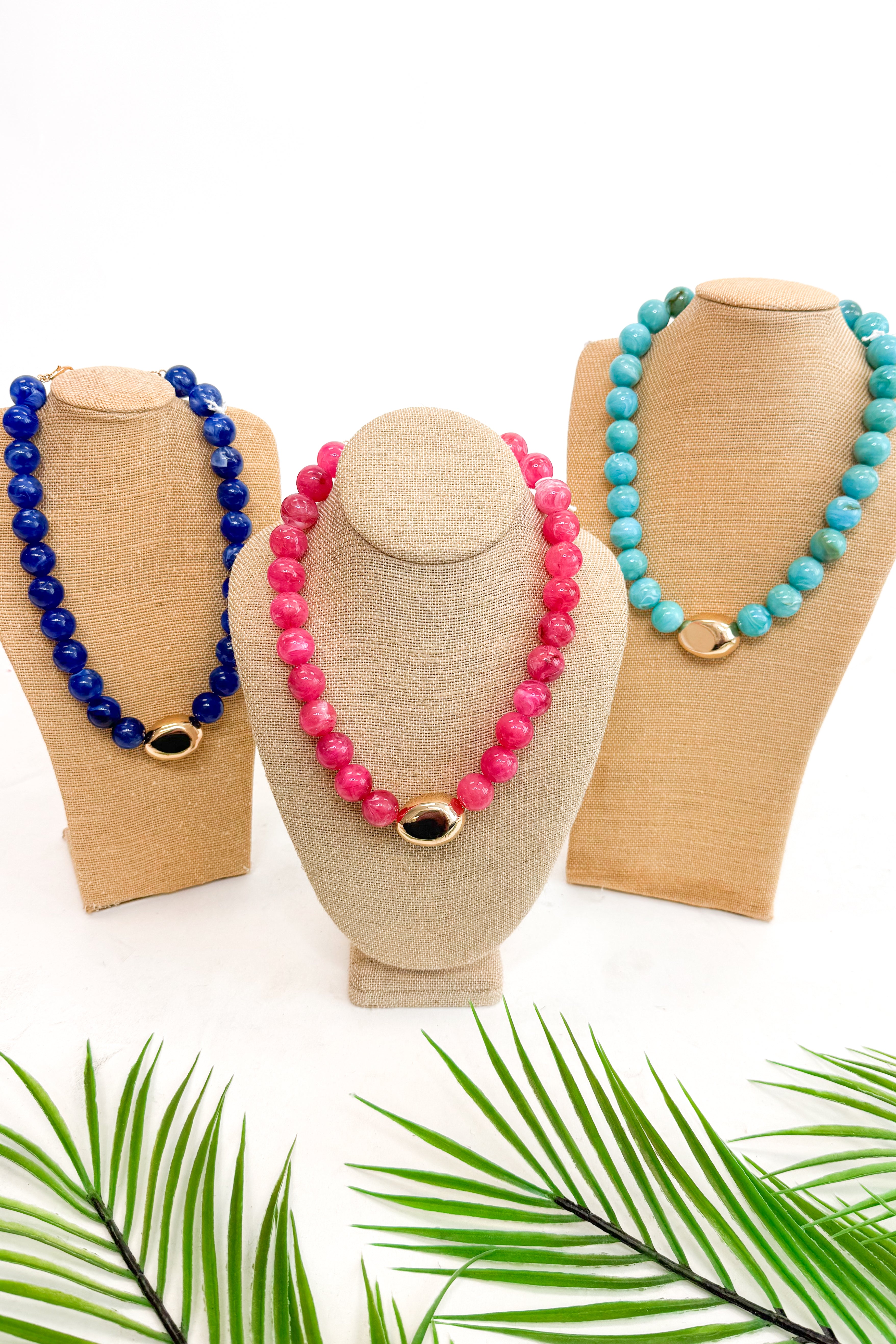 Three beaded necklaces on mannequin heads with a white background and palm leaves at the bottom.