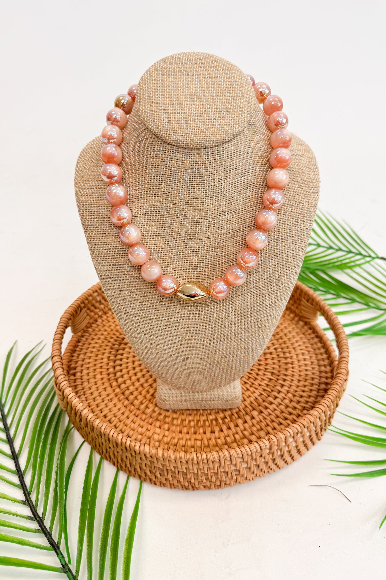 Necklace on a beige display bust with a woven base, surrounded by green leaves.