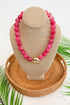 Pink beaded necklace on a beige bust with a woven base, surrounded by green leaves.