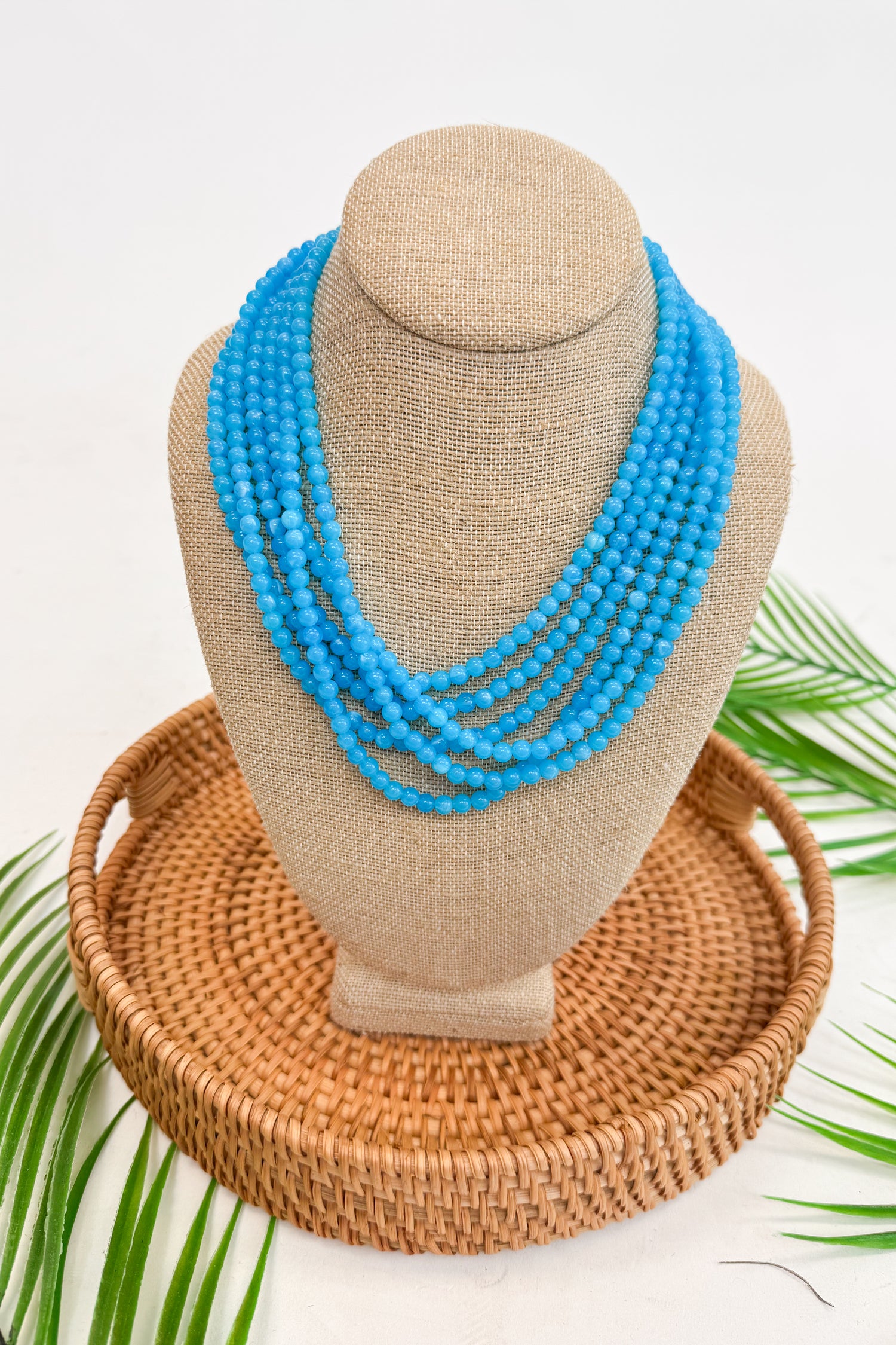Blue beaded necklace on a mannequin bust with a woven basket and palm leaves in the background