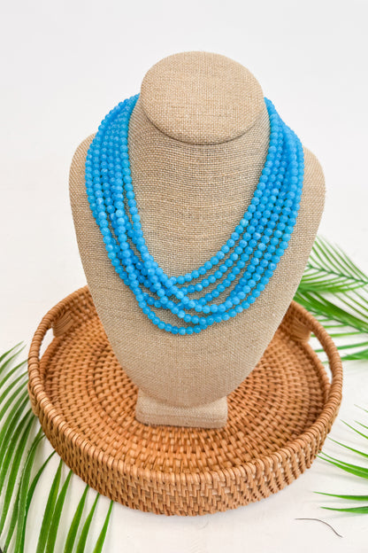 Blue beaded necklace on a mannequin bust with a woven basket and palm leaves in the background