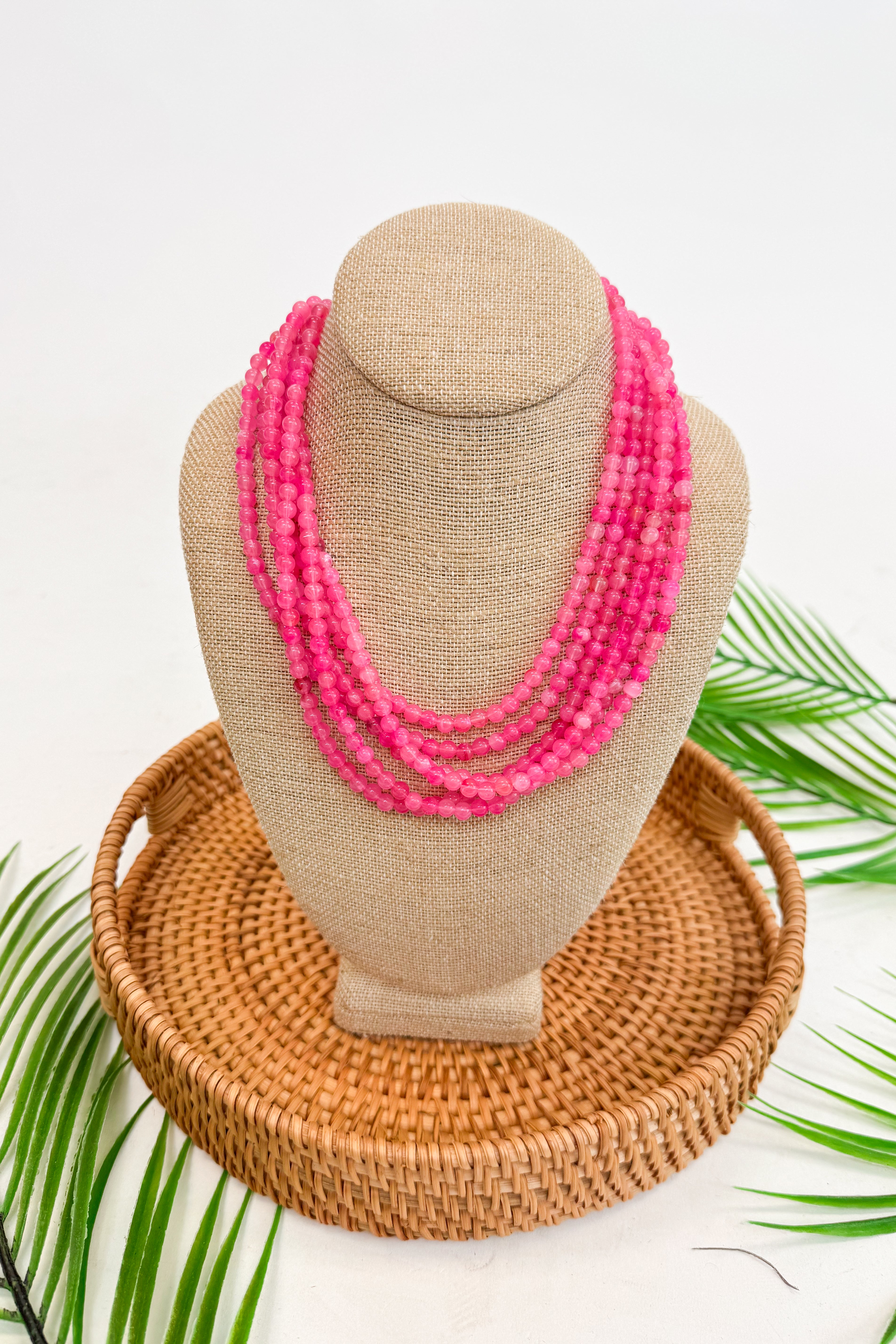 Neon pink beaded necklace on a mannequin bust with a woven basket and palm leaves in the background