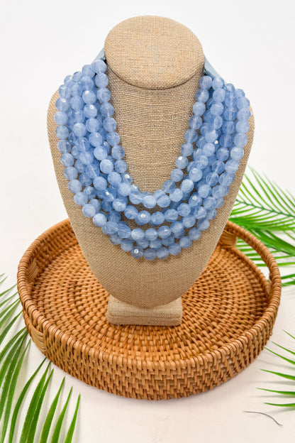 Stack of blue beaded necklaces on a mannequin bust with a woven base and green leaves in the background