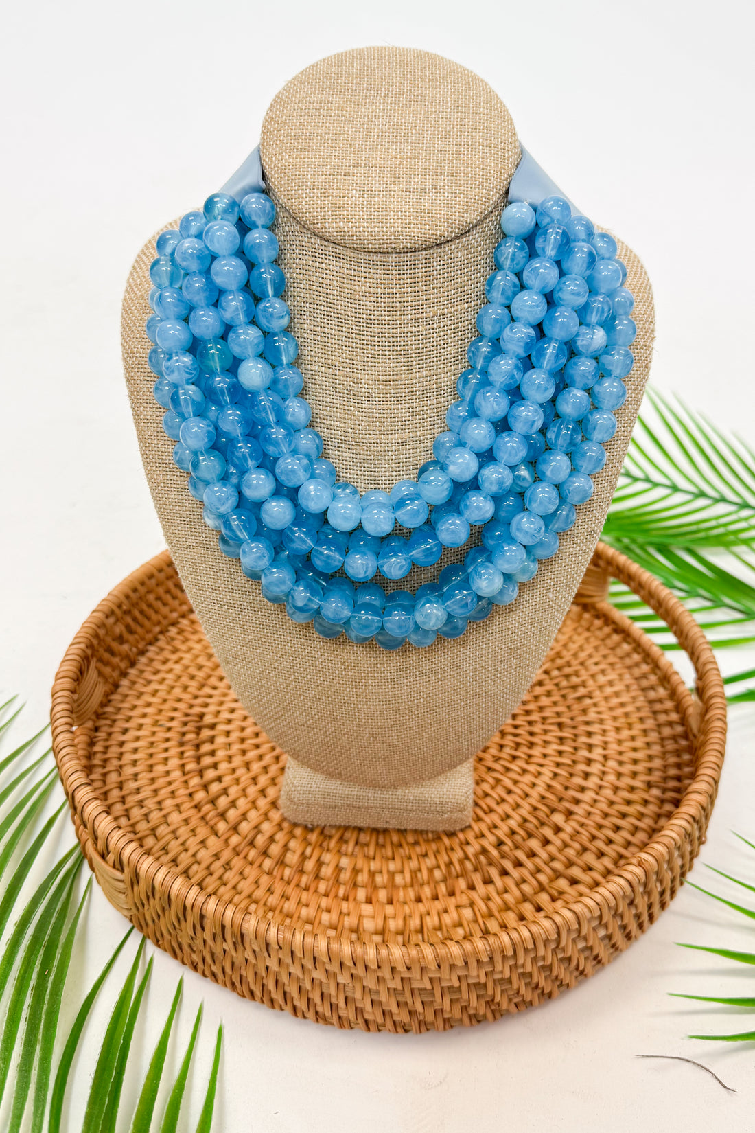 Blue beaded necklace on a tan mannequin bust with a woven base and green leaves in the background