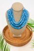 Blue beaded necklace on a tan mannequin bust with a woven base and green leaves in the background