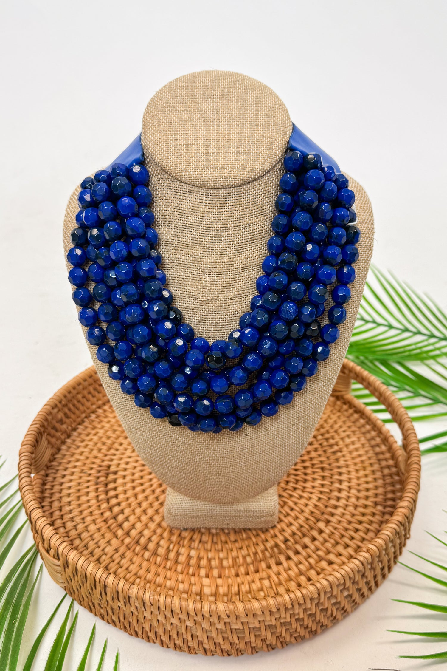 Blue beaded necklace on a mannequin bust with a woven basket and green leaves in the background