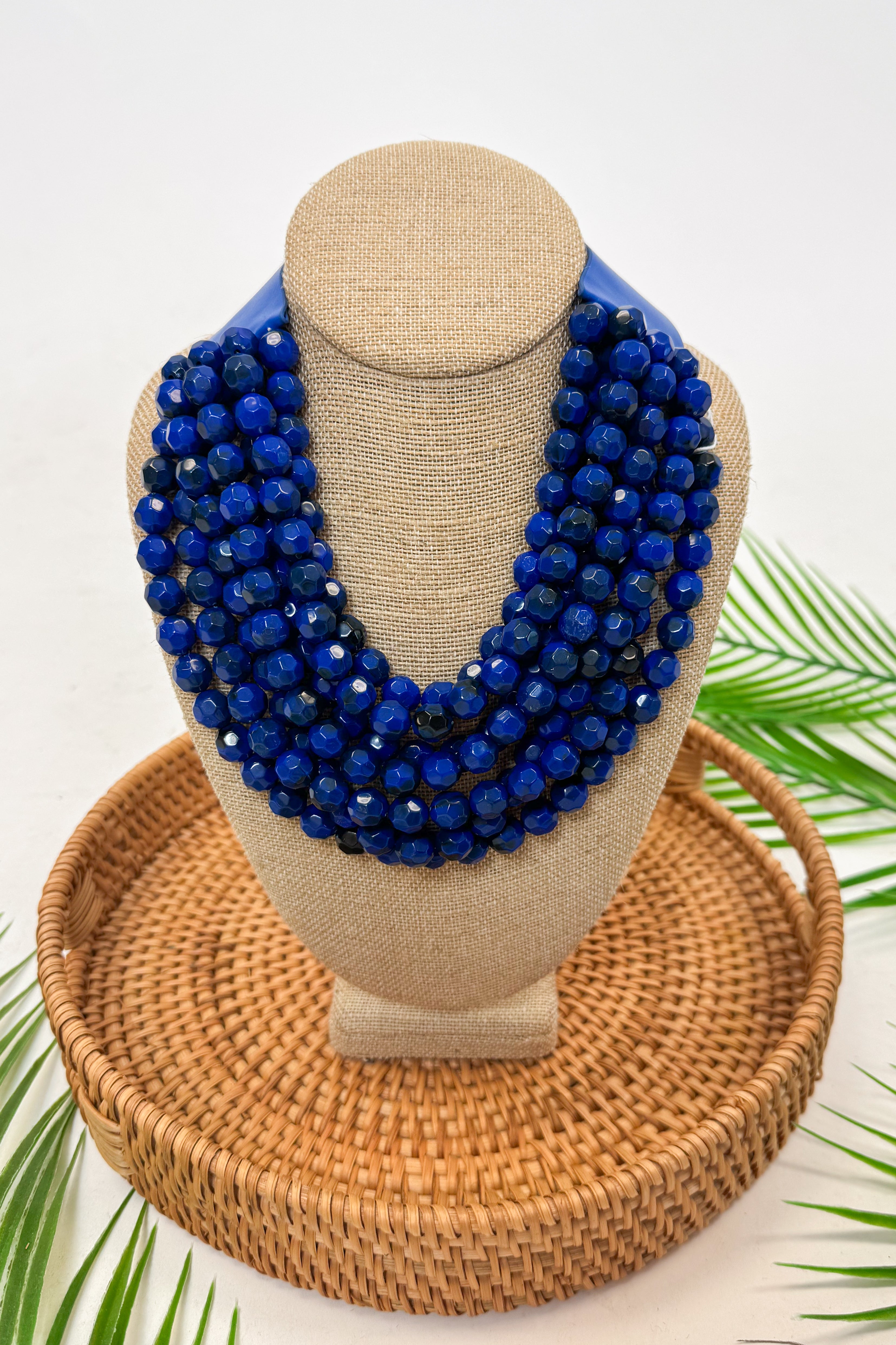 Blue beaded necklace on a mannequin bust with a woven basket and green leaves in the background
