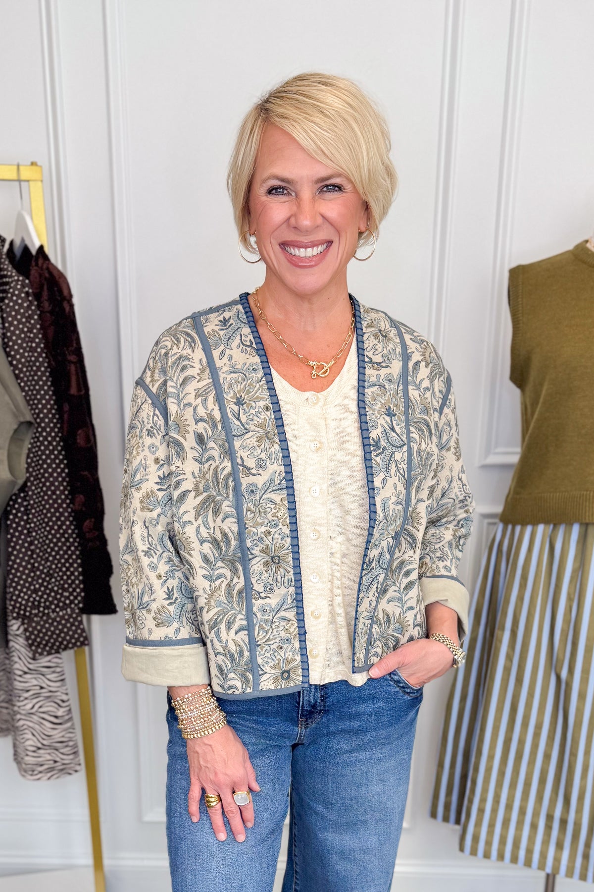 Front view of woman wearing Swagger Boutique’s Beige &amp; Blue Printed Lightweight Jacket, Cream Sleeveless Button up Sweater with Maya Denim Jeans with clothing hanging in the background.