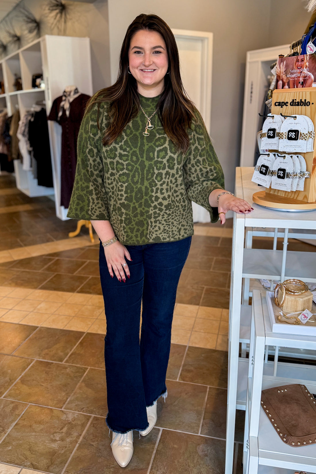 A woman posing in a boutique shop, wearing a green leopard-print long-sleeve top and dark blue flare jeans, standing next to a white display table.