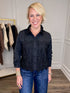 A smiling blonde woman wearing a black long-sleeve button-up blouse with a lace panel across the chest and blue jeans, standing in an indoor boutique setting.